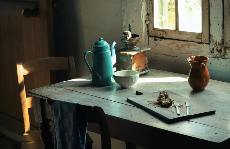 Un tavolo vintage davanti a una finestra con sopra una teiaera di metallo, un macinino da caffè, e alcune stoviglie vintage